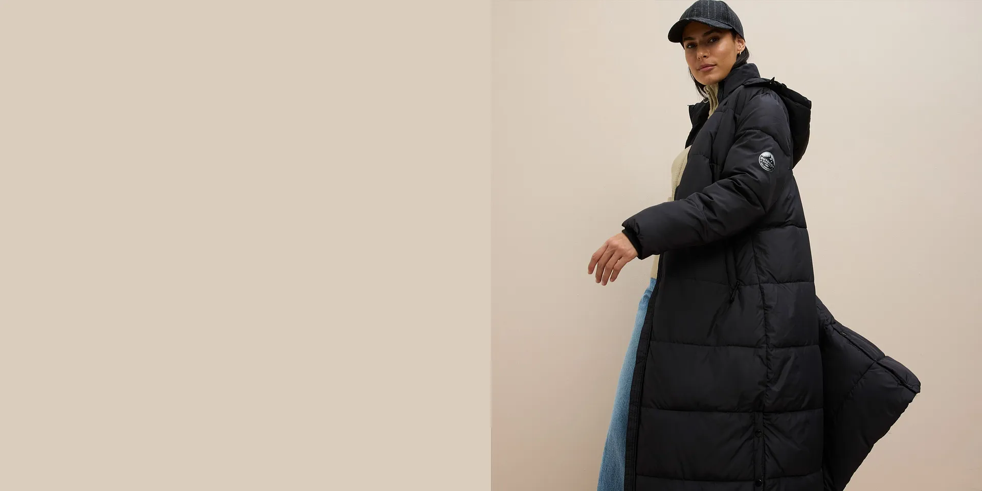 Lange zwarte gewatteerde jas over beige trui, met wijde jas en muts om de look compleet te maken.