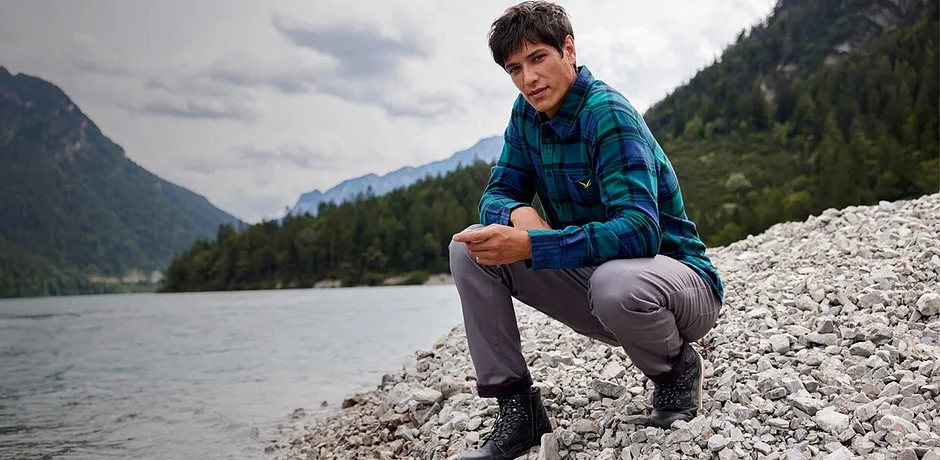 Wandelaar in geruit overhemd en grijze broek zit langs de oever van een rivier in een berglandschap.