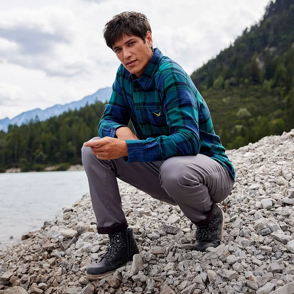 Wandelaar in geruit overhemd en grijze broek zit langs de oever van een rivier in een berglandschap.