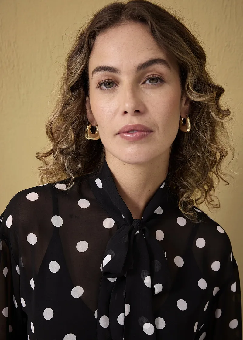 Elegante zwarte blouse met witte stippen en strikkraag. Gouden oorbellen maken de look compleet.