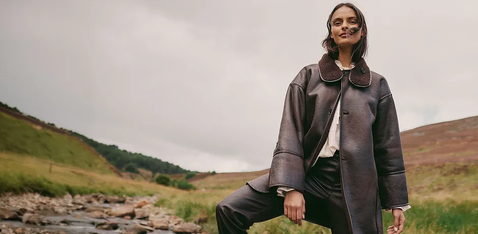 Elegante lange bruine jas van imitatieleer met grote kraag gecombineerd met lichte blouse en donkere broek.