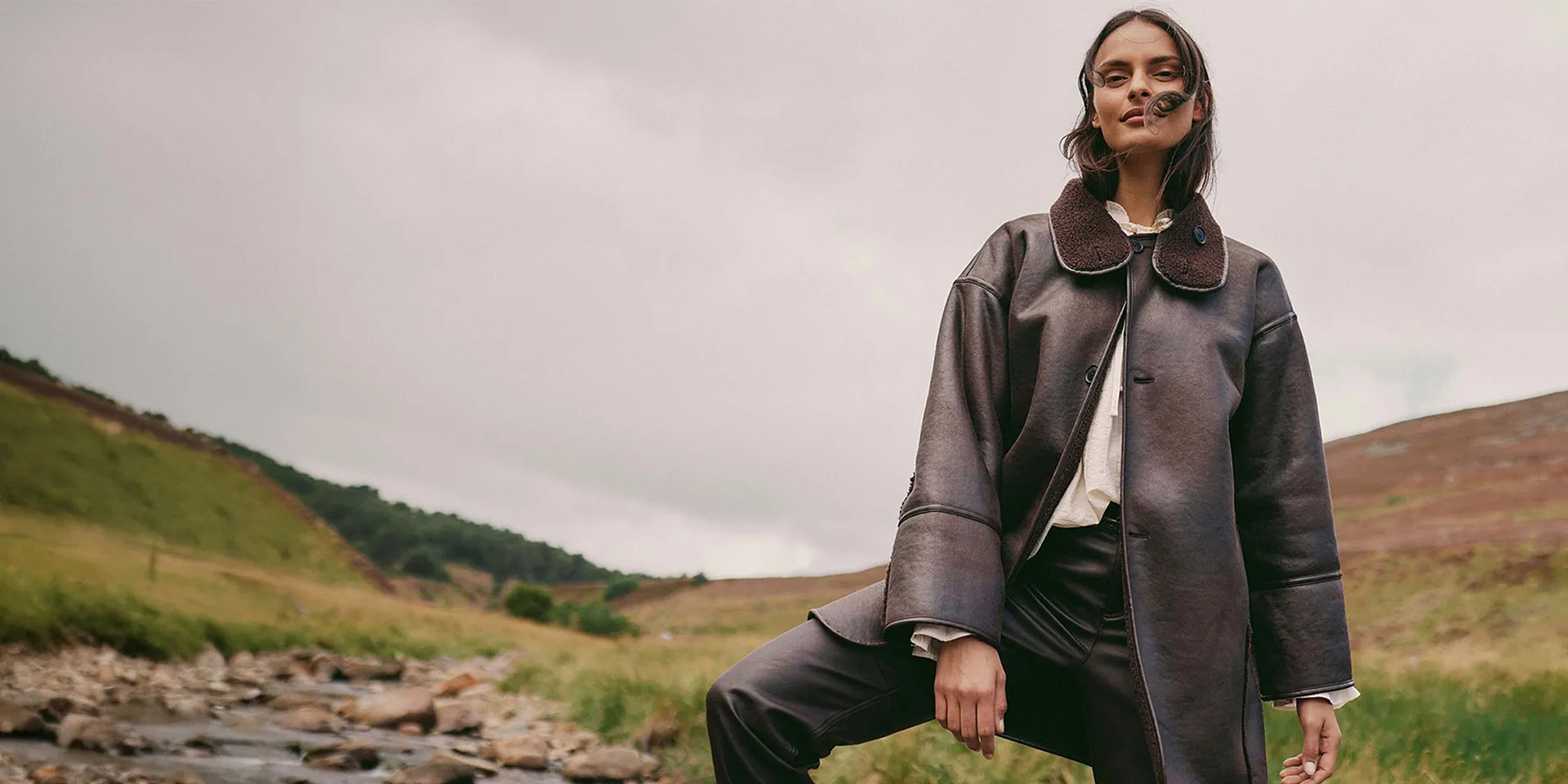 Elegante lange bruine jas van imitatieleer met grote kraag gecombineerd met lichte blouse en donkere broek.