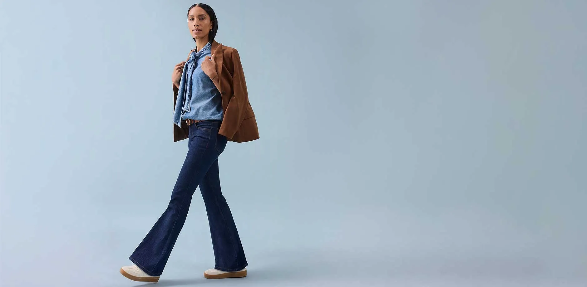 Een vrouw draagt een flared broek, een mouwloos topje en een bruine jas nonchalant over haar schouder.