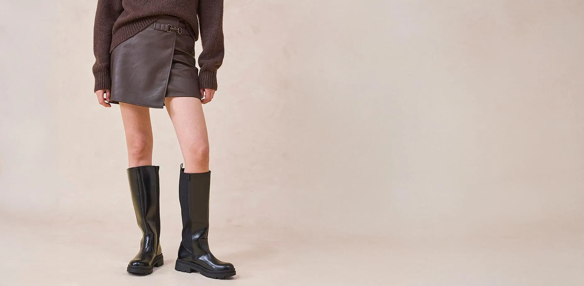 Imitatieleren rok en kniehoge laarzen gecombineerd met een bruine trui.