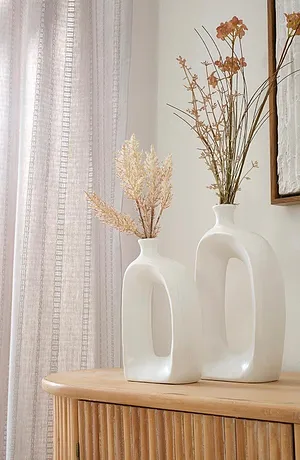 Lichte, luchtige gordijnen omlijsten elegante witte vazen met gedroogde bloemen op een houten kast.