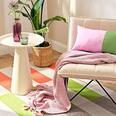 Helle Textilien in Pastellfarben: Rosa Decke, grüne und rosa Kissen auf beigem Sessel.