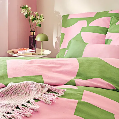 Frisse bedlinnen in roze en groen, gemusterd met geometrische vormen. Een geweven deken complementeert de look.