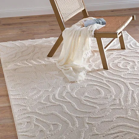 Crème-kleurige deken ligt nonchalant over een rieten stoel op een elegante tapijt met golvende patronen.