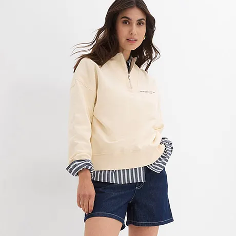 Cremefarbener Pullover über gestreiftem Hemd, kombiniert mit Jeans-Shorts und weißen Sneakers.
