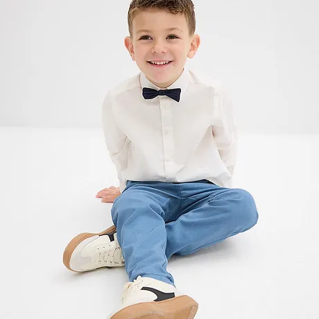 Elegante jongen in een wit shirt met een vlinderdas, lichtblauwe broek en witte schoenen.