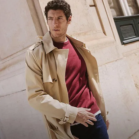 Elegante beigekleurige jas over een rode trui en jeans. Een casual, urban look voor de herfst.