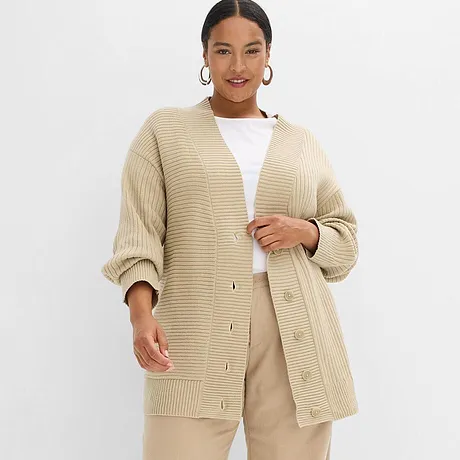 Grof gebreide, beige oversized cardigan gecombineerd met een witte top en een lichtbeige broek.