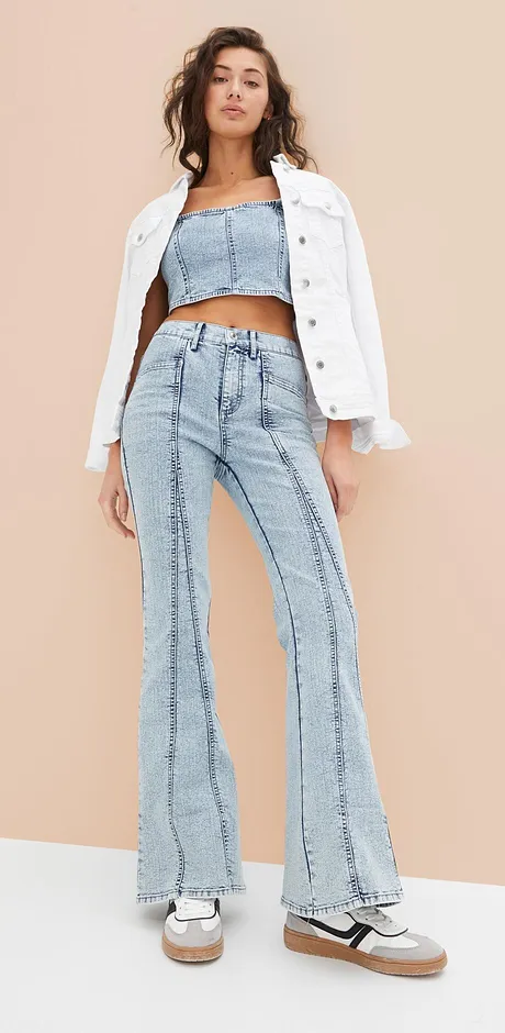 Trendy outfit met een witte spijkerjack, een crop top en een uitlopende spijkerbroek in retro-stijl.