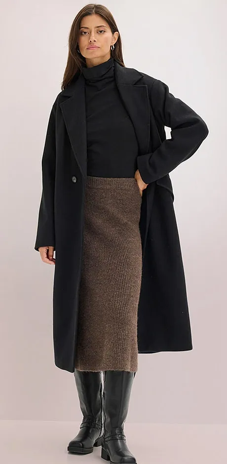 Elegante zwarte jas over coltrui, gecombineerd met bruine midi rok en kniehoge imitatieleren laarzen.