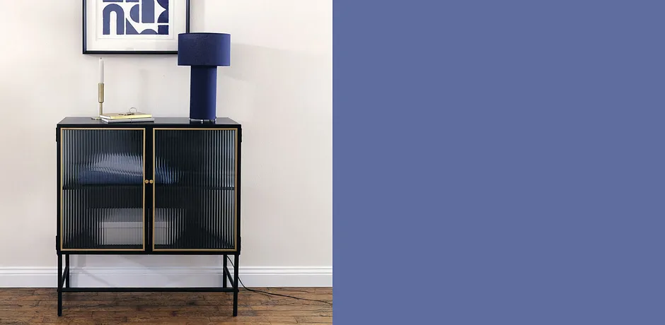 Elegante blauwe tafellamp op een zwarte kast met glazen deuren, daaronder opgevouwen wit textiel.