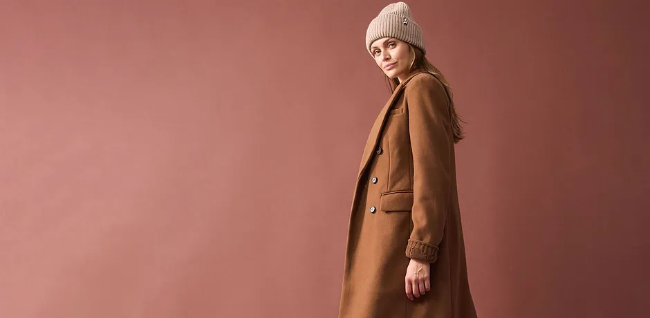 Lange caramelkleurige jas, beige gebreide muts en eenvoudige outfit eronder.